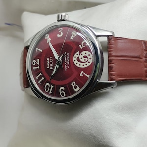 May include: A wristwatch with a round silver-tone case and a deep red dial featuring white Arabic numerals. The dial displays the text HMT PILOT, PARA SHOCK, and 17 JEWELS. It includes a brown leather strap with a textured, embossed pattern.