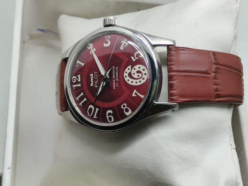 May include: A wristwatch with a round silver-tone case and a deep red dial featuring white Arabic numerals. The dial displays the text HMT PILOT, PARA SHOCK, and 17 JEWELS. It includes a brown leather strap with a textured, embossed pattern.