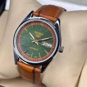 May include: A Seiko 5 automatic watch with a green dial and brown leather strap. The watch face features gold hands and markers, with the day and date displayed. The watch has a stainless steel case and a brown leather band.