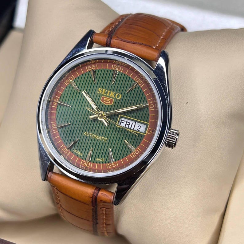May include: A Seiko 5 automatic watch with a green dial and brown leather strap. The watch face features gold hands and markers, with the day and date displayed. The watch has a stainless steel case and a brown leather band.