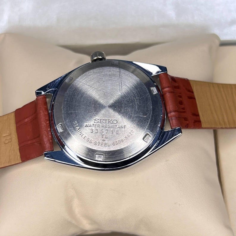 May include: A stainless steel Seiko watch with a red leather band. The watch face is silver with the words "SEIKO WATER RESISTANT STAINLESS STEEL" and other text. The watch band is a reddish-brown color.