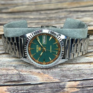 May include: A silver-toned Seiko automatic wristwatch with a green dial and a stainless steel band. The watch face displays the day of the week as "MON 1" and the brand name "SEIKO". The watch is resting on a gray surface.
