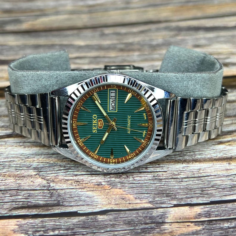 May include: A silver-toned Seiko automatic wristwatch with a green dial and a stainless steel band. The watch face displays the day of the week as "MON 1" and the brand name "SEIKO". The watch is resting on a gray surface.