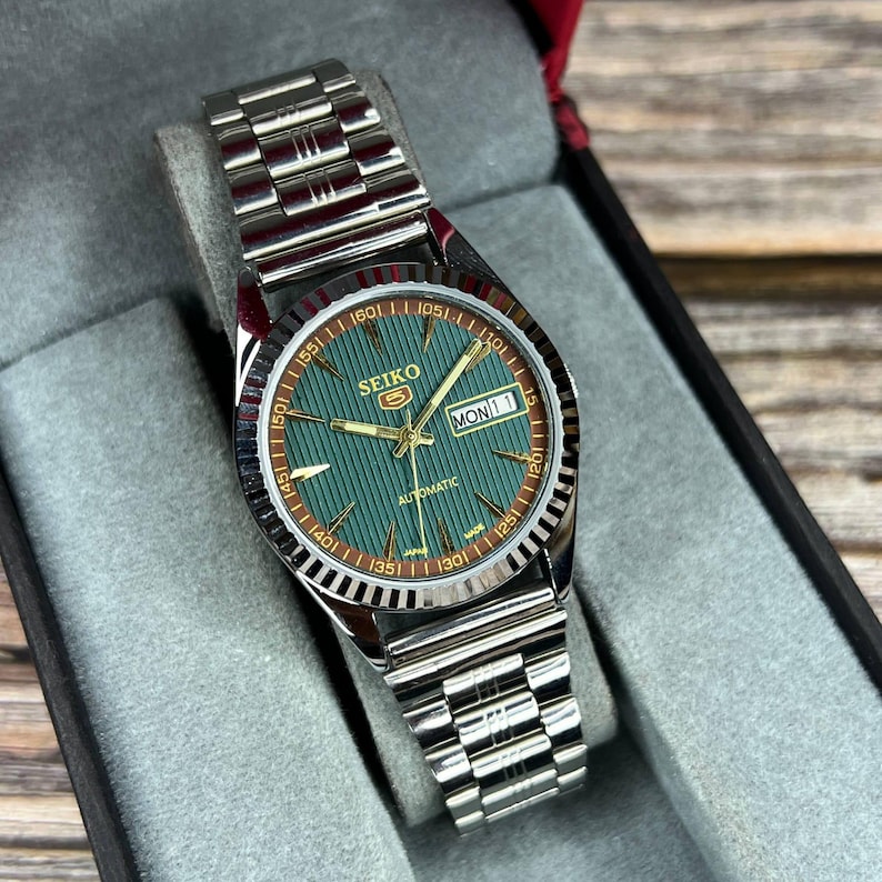 May include: A silver-toned Seiko automatic wristwatch with a green dial and a silver band. The watch face displays the day and date, with gold-colored hands and markers. The watch is presented in a gray velvet-lined box.