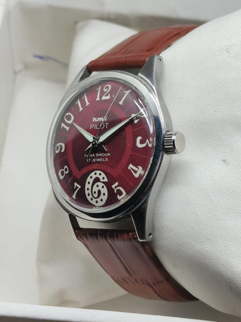 May include: A wristwatch with a silver-tone circular case and a deep red dial featuring large white Arabic numerals. The dial displays the text HMT PILOT, PARA SHOCK, 17 JEWELS, and INDIA. It includes a brown leather strap with a textured finish.