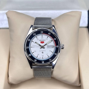 May include: A silver Seiko automatic watch with a white face, black and red details, and a mesh strap. The watch shows the date as Saturday the 26th. The watch is displayed on a cream cushion inside a box.