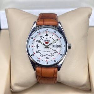May include: A silver-toned Seiko automatic watch with a white face and brown leather strap. The watch face displays the day and date, with black numerals and markers. The watch is presented on a beige pillow.
