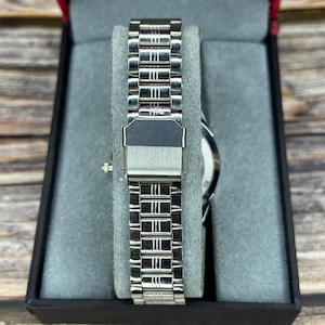 May include: A silver-toned wristwatch with a stainless steel band is displayed in a black box with a gray interior. The watch band is composed of rectangular links. The watch face is visible.