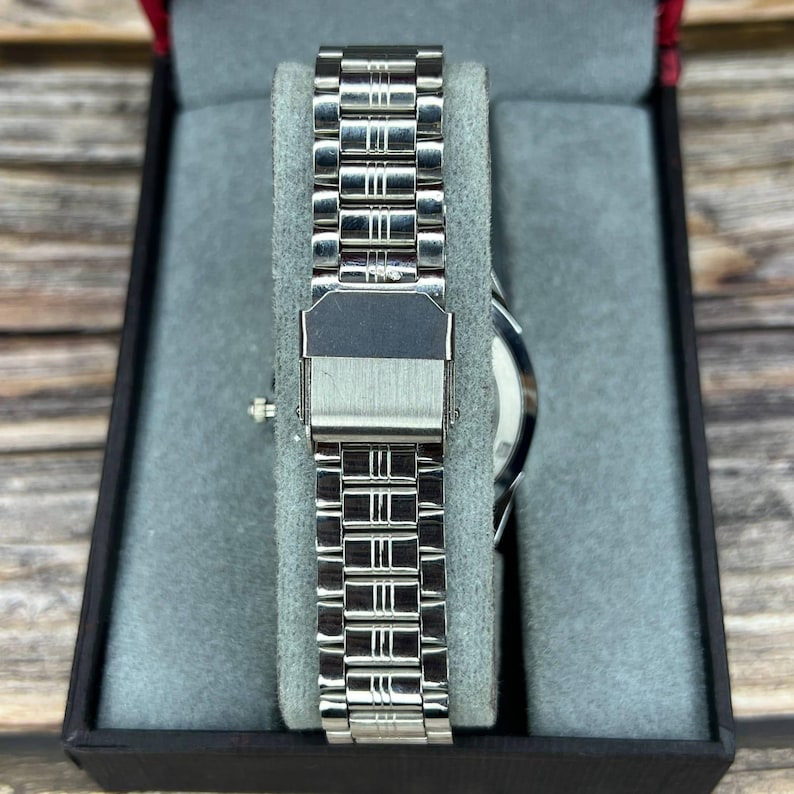 May include: A silver-toned wristwatch with a stainless steel band is displayed in a black box with a gray interior. The watch band is composed of rectangular links. The watch face is visible.
