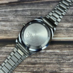 May include: A stainless steel Seiko watch, viewed from the back, with a silver metal band. The watch face is silver with black text that reads "SEIKO WATER RESISTANT 335718 TL 6300". The watch is on a wooden surface.