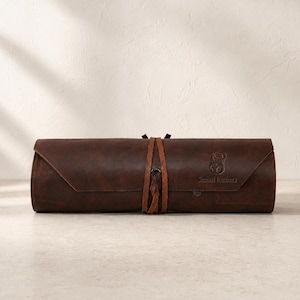 May include: A dark brown leather roll-up case featuring a wrap-around tie closure. The exterior is embossed with a bull head logo and the text "Sanaal leathers" on the right side. The case has a smooth, matte finish and a structured cylindrical shape.