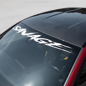 May include: A white "SAVAGE" decal is affixed to the windshield of a car. The decal is in a stylized font with sharp edges and a trailing design. The car has a black roof and a red body.