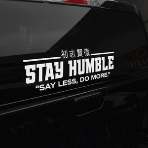 May include: White vinyl decal with the words "STAY HUMBLE" in bold, block letters, and the phrase "SAY LESS, DO MORE." The decal also includes Japanese characters. The sticker is affixed to the black window of a vehicle.