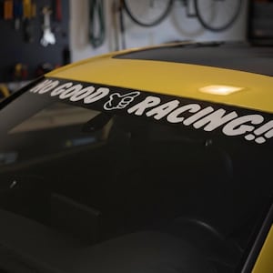 May include: A yellow car with a black windshield. A white decal across the top of the windshield reads "NO GOOD RACING!!" in a bold, stylized font. The car is parked in a garage.