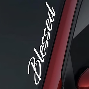 May include: White vinyl decal with the word "Blessed" in a cursive font. The decal is vertically oriented and placed on a car window, with a red car door visible in the background.