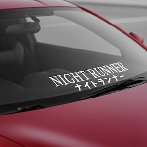 May include: A close-up of a red car's windshield with a white decal. The decal reads "NIGHT RUNNER" in a bold, sans-serif font, with Japanese characters below. The car's interior is visible through the glass.
