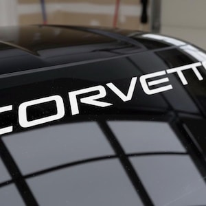 May include: A black car with a white "CORVETTE" decal across the top of the windshield. The decal is in a bold, sans-serif font. The car's glossy surface reflects the surrounding environment.