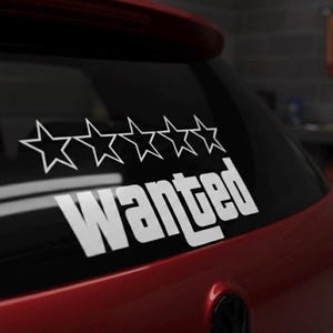 Puede incluir: Una calcomanía blanca de "Wanted" con cinco contornos de estrellas está adherida a la ventana trasera de un coche rojo. El texto y las estrellas están en una fuente en negrita sin serifa.