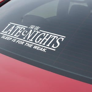 May include: White vinyl decal on a car window. The decal reads "LATE NIGHTS" with Japanese characters above and "SLEEP IS FOR THE WEAK." below. The car is red.