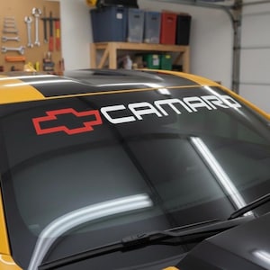 Puede incluir: Coche deportivo amarillo con capó negro y una pancarta en el parabrisas que dice "CAMARO" en letras blancas. Un logotipo de Chevrolet rojo y blanco está a la izquierda. El coche está en un garaje.