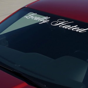 Puede incluir: Primer plano del parabrisas de un coche rojo con una calcomanía blanca que dice "Locally Hated" en una fuente cursiva. El espejo lateral del coche es visible, y el fondo es una superficie gris claro.