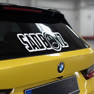 May include: A yellow BMW car with a black rear window. A white sticker with the text "SADBOI" and a sad face graphic is on the window. The BMW logo is visible.