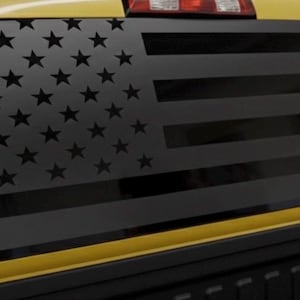 May include: Black and grey American flag decal on the rear window of a yellow truck. The flag design features black stars and stripes, creating a patriotic appearance. The decal is a decorative accessory for vehicles.