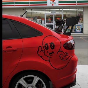 Puede incluir: Coche rojo con portón trasero, alerón negro y un dibujo animado en el lateral. El dibujo es una figura fantasmal haciendo una peineta. El coche está aparcado frente a una tienda 7-Eleven.