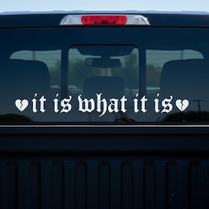 May include: White vinyl decal applied to a vehicle's rear window. The decal features the phrase "it is what it is" in a gothic font, with a broken heart symbol on each side of the text. The vehicle's black tailgate is visible.