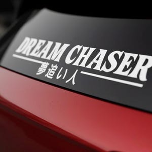 May include: White "DREAM CHASER" decal with Japanese characters, applied to a black surface. The decal is on a red car. The text is in a bold, sans-serif font.