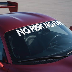 Puede incluir: Un coche deportivo granate con una pancarta blanca en el parabrisas. La pancarta dice "NO RISK NO FUN" en letras negritas y estilizadas. El coche tiene un alerón y espejos laterales rojos. El interior del coche es visible.