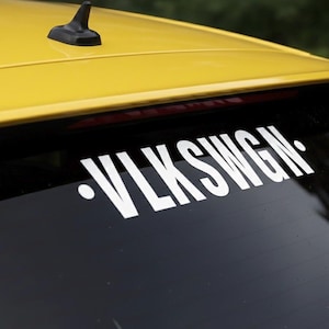 Puede incluir: Un coche amarillo con una luneta trasera negra que muestra una calcomanía blanca con la inscripción "VLKSWGN". El coche tiene una antena negra y una luz de freno roja. La calcomanía está centrada en la ventana.