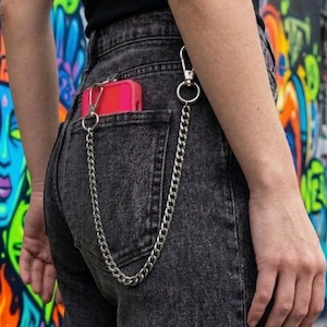 Puede incluir: Vaqueros de mezclilla gris oscuro con una cadena plateada sujeta al bolsillo trasero. Se ve una funda roja para teléfono en el bolsillo. El fondo presenta arte de graffiti colorido.