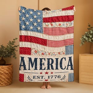 May include: A rectangular quilt featuring a patchwork design of the American flag with various red, white, and blue fabric patterns. The word AMERICA is printed in large dark blue letters above the text EST. 1776, accented by three small red stars.