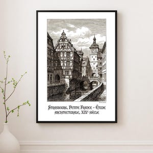 May include: Black and white framed print of Strasbourg, Petite France. The architectural study from the 19th century depicts buildings along a canal with a bridge and a clock tower. The print is displayed on a white wall.