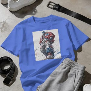 Op de afbeelding: Een blauw t-shirt met korte mouwen en een centrale grafische print van een robotfiguur in een rood jasje en een witte broek. De robot is afgebeeld vanuit een laag standpunt met blauwe en rode accenten op een lichte achtergrond.