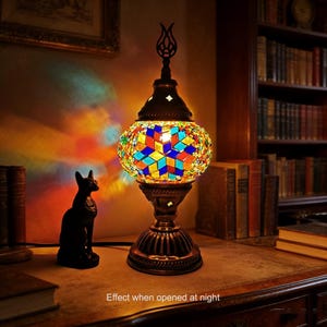 May include: A decorative table lamp with a mosaic glass shade in a variety of colors, including blue, yellow, and red. The lamp has a dark metal base and a decorative top. The text "Effect when opened at night" is visible at the bottom.