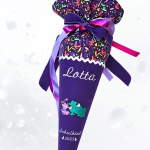 Könnte beinhalten: Eine lila Schultüte mit dem Namen "Lotta" in weißer Schrift. Die Tüte zeigt ein Frosch- und Blumenmuster mit dem Text "Schulkind 2025". Sie ist mit lila und rosa Bändern und einer Schleife gebunden und hat eine floral gemusterte Oberseite.