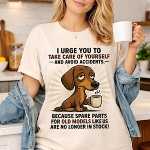 May include: Cream-colored t-shirt featuring a cartoon dachshund dog holding a mug. The shirt has the text: "I URGE YOU TO TAKE CARE OF YOURSELF - AND AVOID ACCIDENTS. BECAUSE SPARE PARTS FOR OLD MODELS LIKE US ARE NO LONGER IN STOCK!"