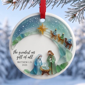 Puede incluir: Un adorno navideño redondo de cerámica blanca con una escena de natividad de vidrio marino, el texto "the greatest gift of all", "MATTHEW 1:21" y "2025". El diseño incluye una estrella dorada, tres camellos y figuras en tonos azules y verdes.