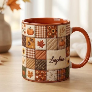 Könnte beinhalten: Ein Keramikbecher mit Patchwork-Design, das herbstliche Quadrate mit Kürbissen, Ahornblättern, Sonnenblumen, Eicheln und braunen Vichy-Karos zeigt. Der Name Sophia ist in dunkelbrauner Schreibschrift auf einem cremefarbenen Quadrat gedruckt.
