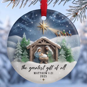 Puede incluir: Un adorno navideño redondo de cerámica con una escena de nacimiento, un establo de madera, figuras y pinos verdes bajo un cielo estrellado. Incluye una estrella dorada, tres camellos y el texto: the greatest gift of all MATTHEW 1:21 2025.