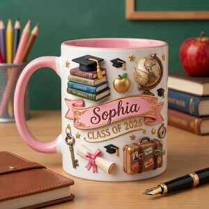 Könnte beinhalten: Weiße Keramiktasse mit rosa Griff und Innenrand. Die Tasse ist mit Elementen zum Thema Abschluss verziert, darunter Bücher, eine Doktorhut, ein Globus und ein Banner mit der Aufschrift "Sophia CLASS OF 2026".