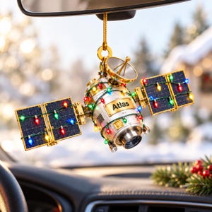 Op de afbeelding: Een satellietvormig hangend ornament met een zilverkleurig cilindrisch lichaam met het woord Atlas op een gouden plaatje. De satelliet heeft twee zonnepanelen versierd met veelkleurige kerstverlichting en een gouden lus om op te hangen.