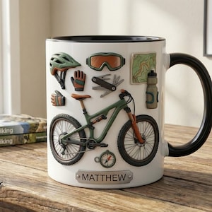 May include: White ceramic mug with a black handle and rim, decorated with mountain biking gear. The design includes a green mountain bike, helmet, goggles, gloves, map, and a water bottle. The name "MATTHEW" is displayed on the mug.
