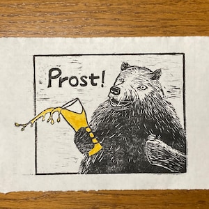 May include: A black and white linocut print showing a bear holding a beer glass, with the word "Prost!" above. The bear has a surprised expression, and the beer is yellow. The print is on textured paper.