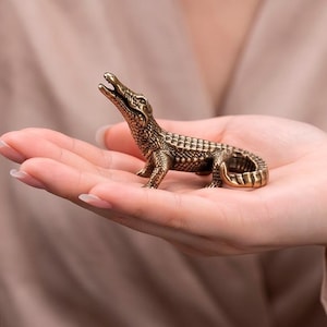May include: A small, detailed brass alligator figurine with an open mouth, held in a hand. The alligator has textured scales and is posed with its head raised. The figurine is a decorative item.