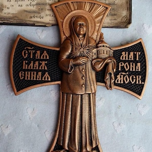 May include: A bronze-colored religious cross featuring a figure holding a building and a cloth. The cross has black accents and Cyrillic text on the arms. An open book with handwritten text is in the background.