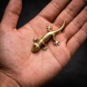 May include: A small, golden lizard figurine with detailed legs, a long tail, and a textured body. The lizard is resting on a palm, with a dark background. The figurine is made of metal and has a polished finish.