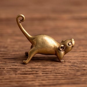 May include: A small, metallic gold-toned brass figurine of a mouse in a stretching pose. The mouse has a long, slender tail that curls at the tip, pointed ears, and a smooth, polished surface. The figure is captured in a dynamic, arched posture.